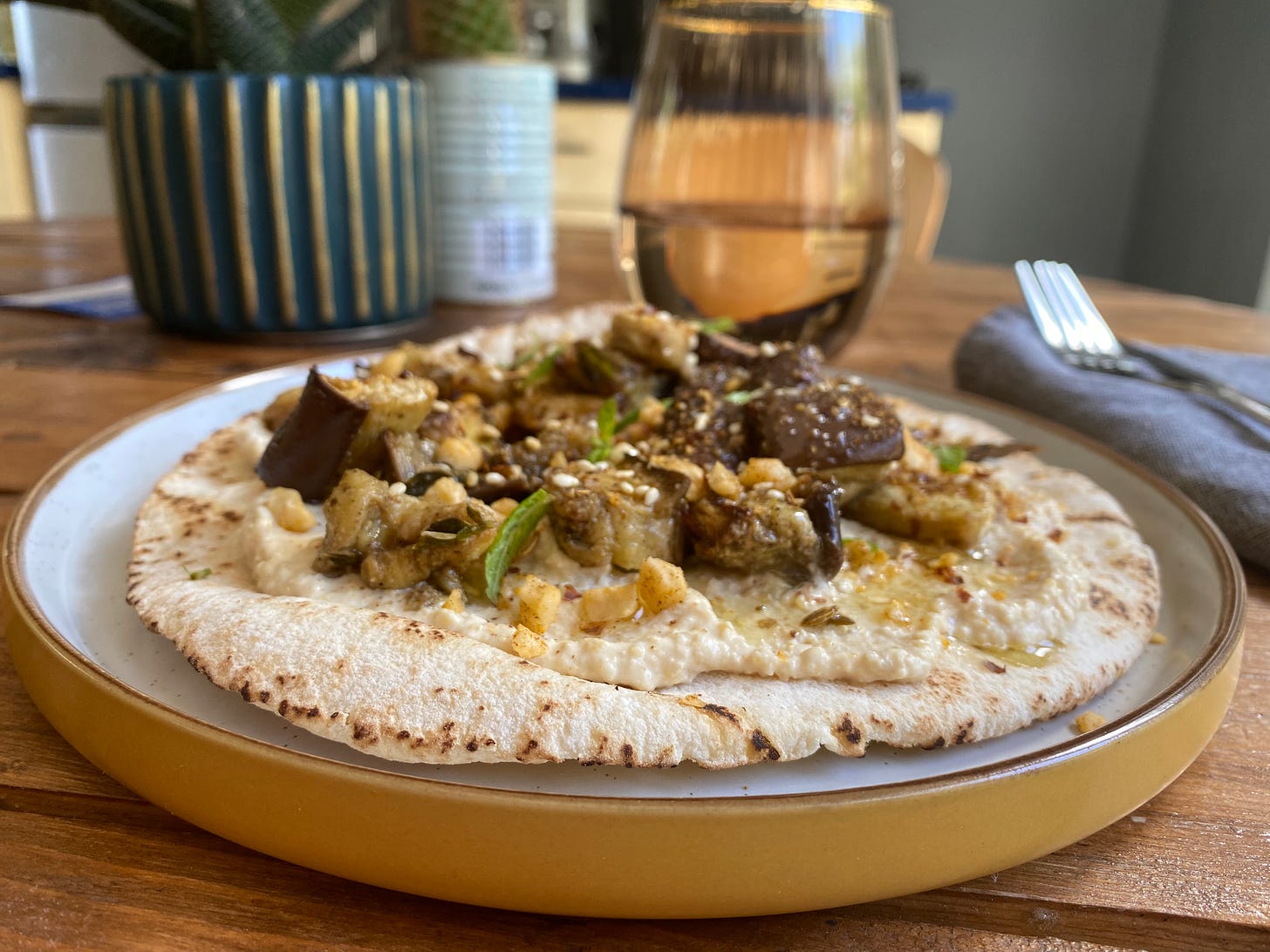 Open flatbread with houmous, aubergine, mint and toasted nuts and seeds. Flatbread is on a yellow-rimmed plate on a wooden table. A glass, and cutlery are placed nearby.