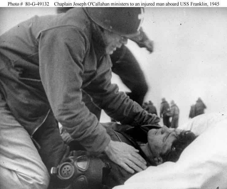 Photo of Father O'Callahan tending to an injured man aboard USS Franklin. O'Callahan has his hands on the shoulders of the injured man, who is lying on the ground.