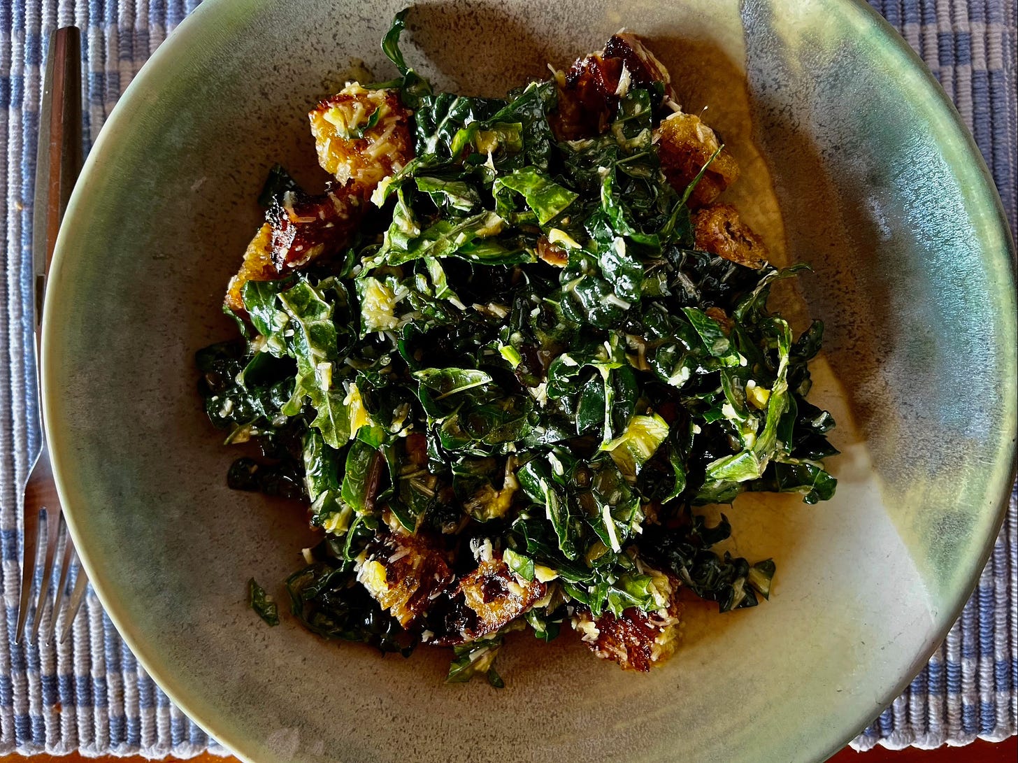 A green and white ceramic bowl of kale Caesar salad: roughly chopped bright green leaves and croutons, with lots of pressed garlic and Parmesan visible on top.