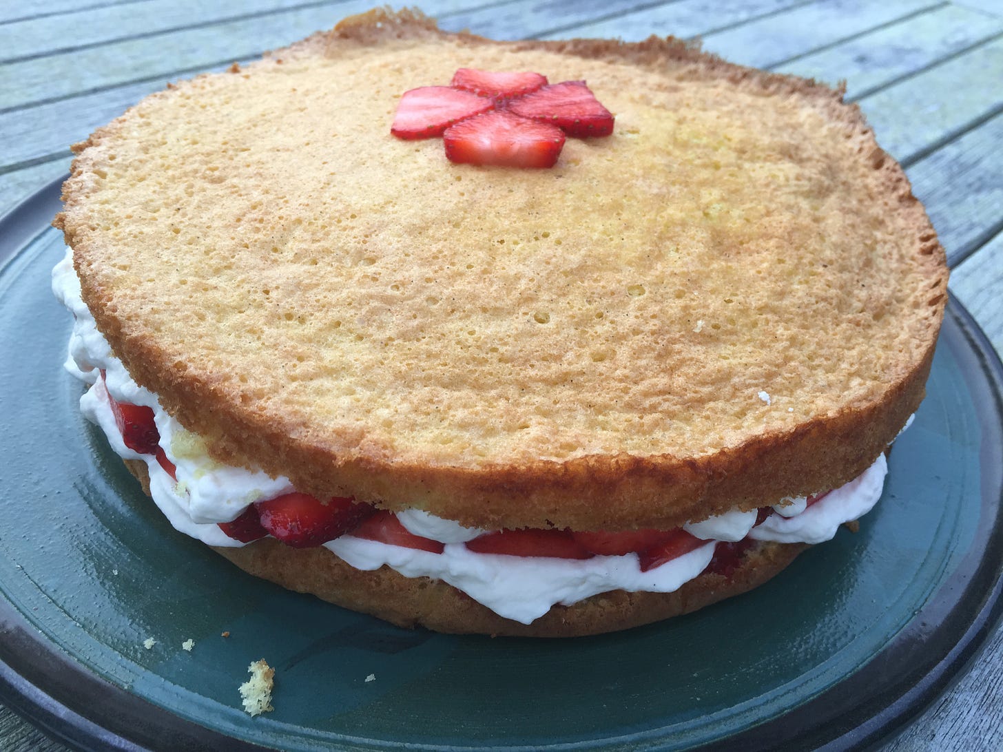 A large golden two-layer cake sits on a ceramic plate. It is filled with sliced strawberries and whipped cream, which is oozing out the sides.