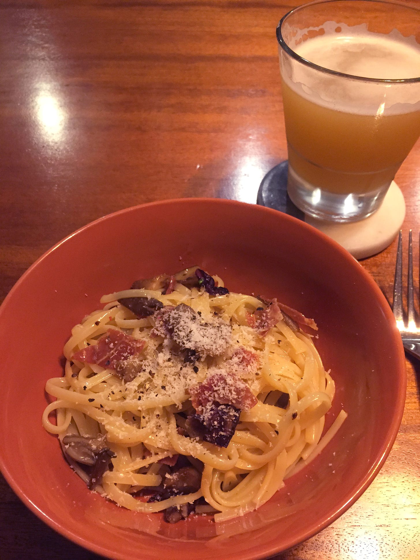 An orange bowl of linguine carbonara with dark purple pieces of radicchio and chopped oyster mushrooms visible throughout. Pecorino and ground pepper are dusted over the top. A glass of hazy IPA sits above and to the right of the bowl on a coaster.