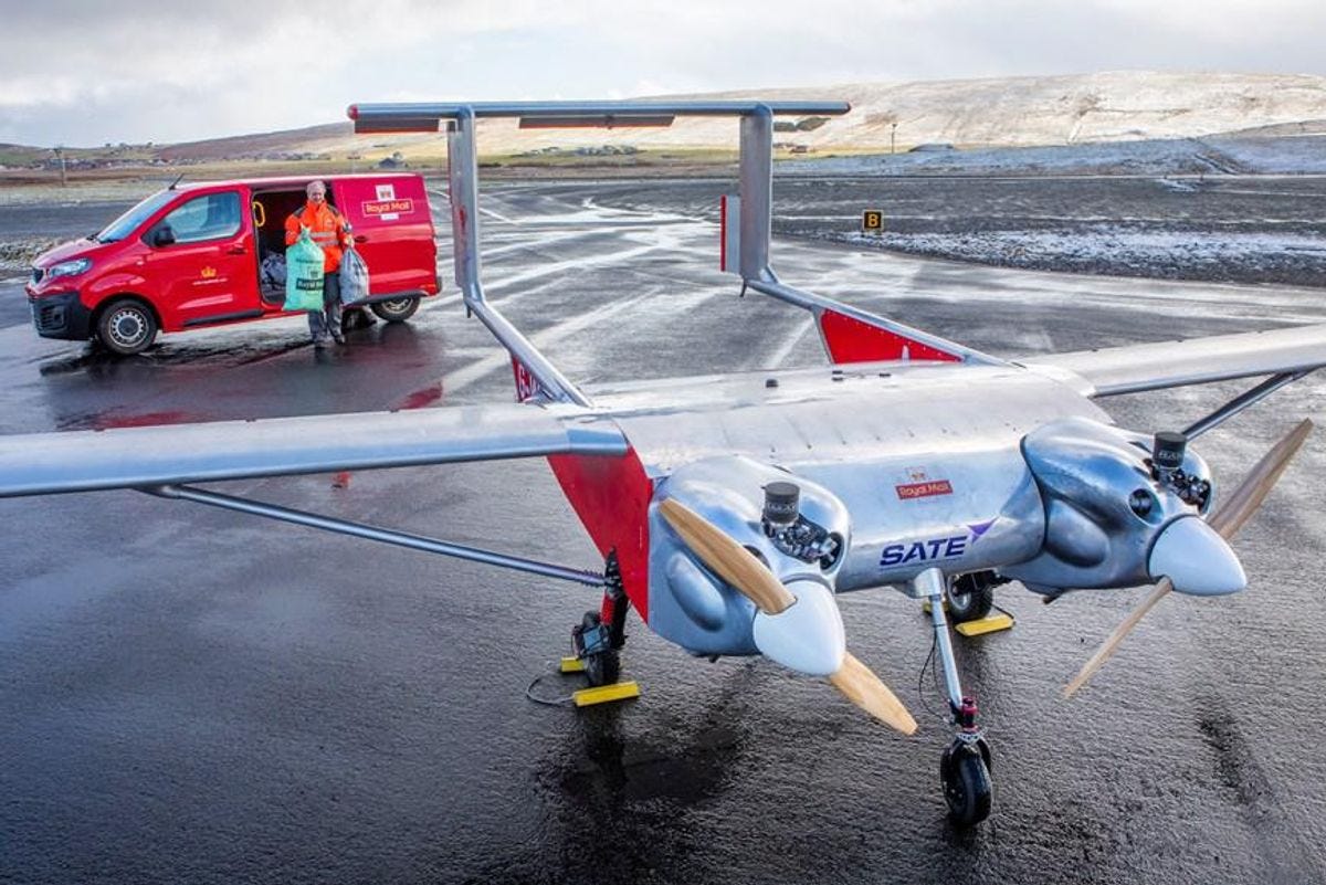A large drone with twin propellers stands idle on a remote airport runway as a postal worker walks towards it with two large mail bags