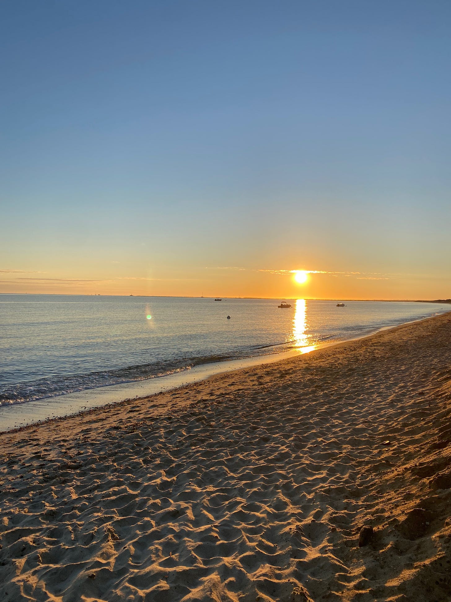 A beach at sunrise. The golden sun is low on the horizon, casting a golden trail across the still water. The beach is sunlit and the sky is pale blue. 