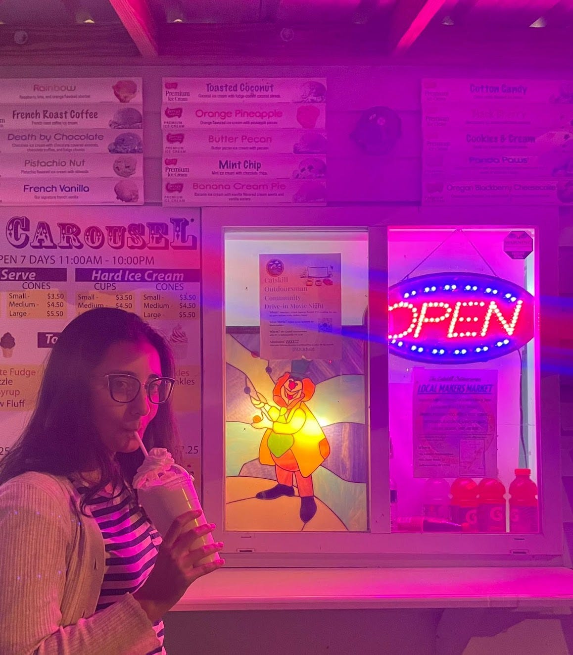 A young woman with black hair and glasses drinks a milkshake in front of an ice cream stand