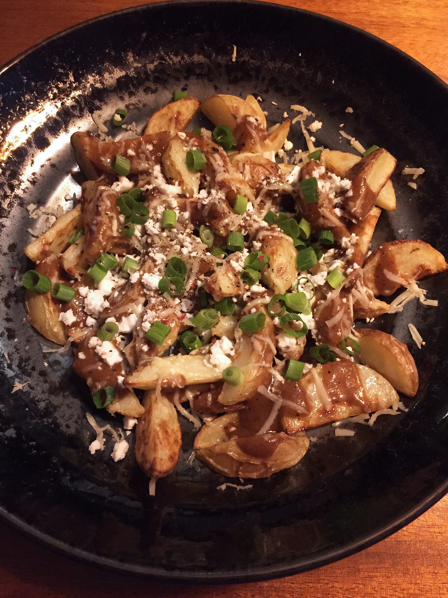 a black bowl filled with small wedge fries and covered in gravy, shredded mozzarella, and crumbles of feta, with chopped green onions.