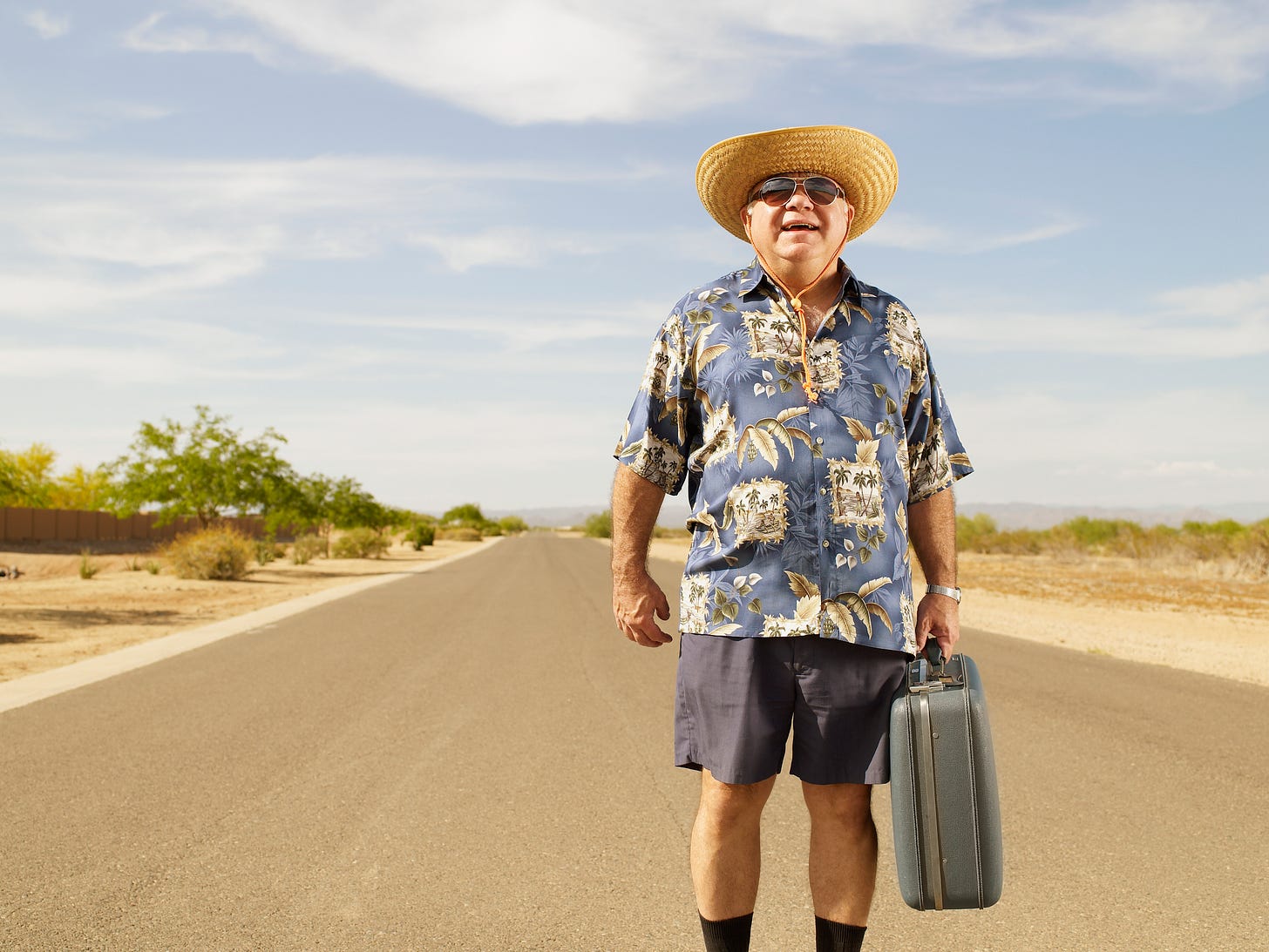 Man in straw hat and Hawaiian style shirt holding briefcase in the middle of an empty highway
