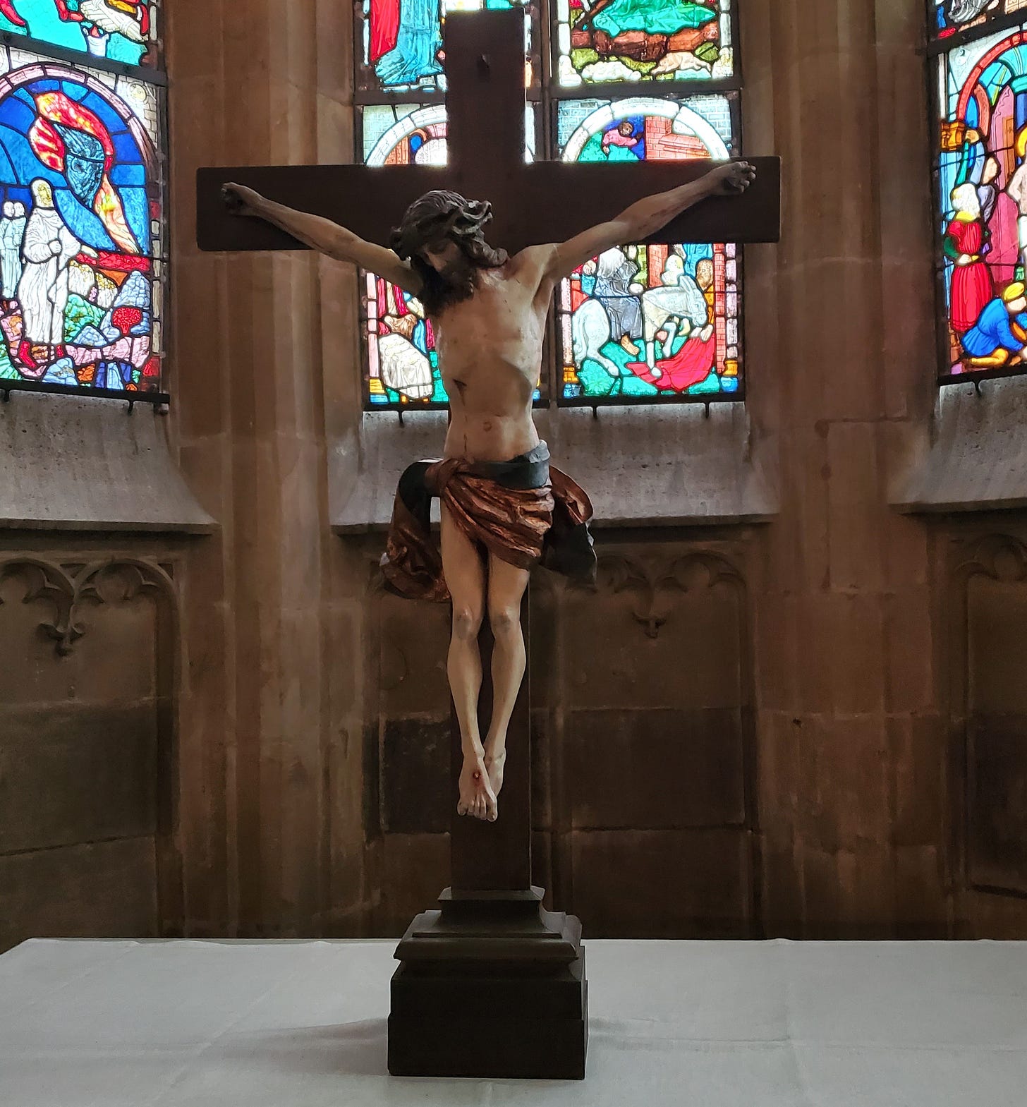 A not-very bloody crucifix in front of some stained glass windows.