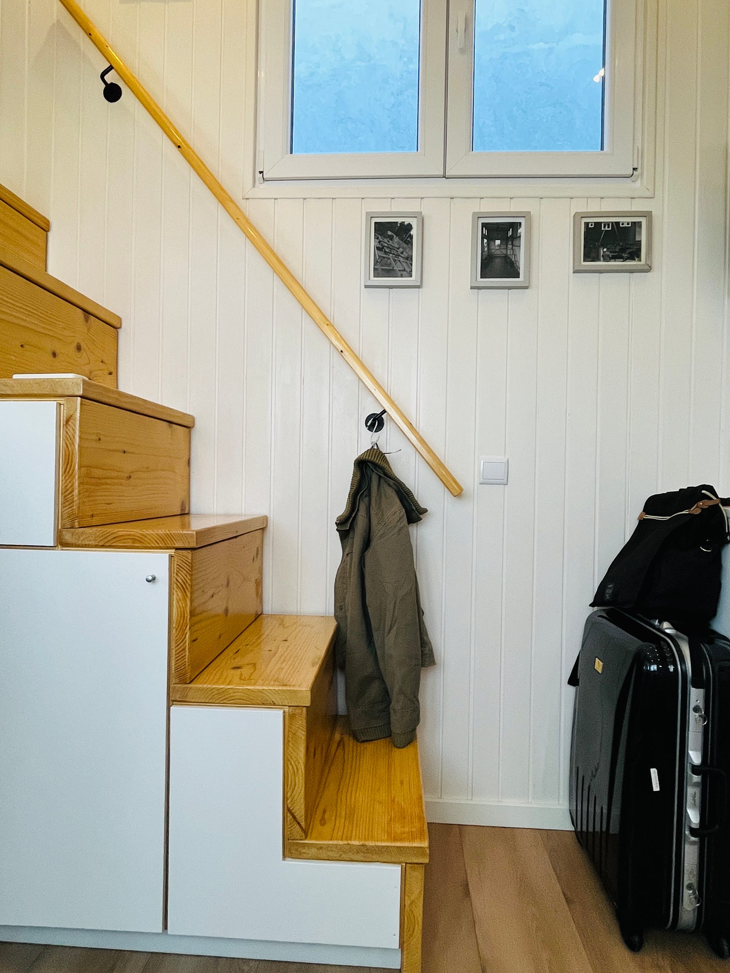 Image: photo of a wooden staircase, a jacket hanging from the staircase rail, two white windows on the top, and on the right, a black colour suitcase and a black colour backpack.