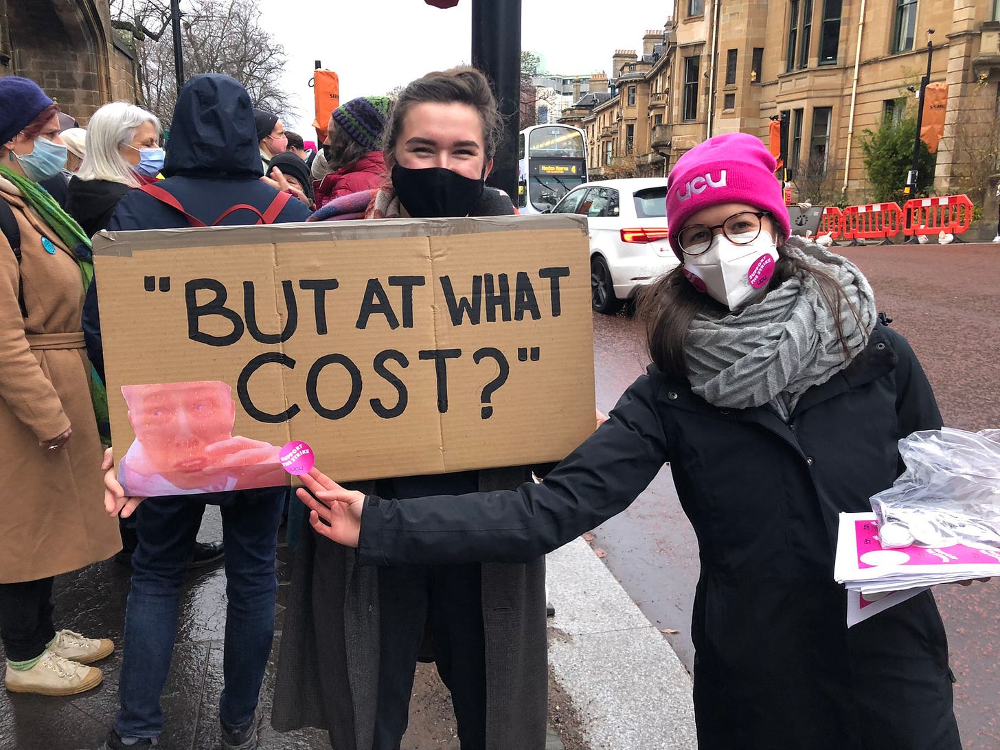 ID: A sign being held up by two protesters that says: "At what cost?"