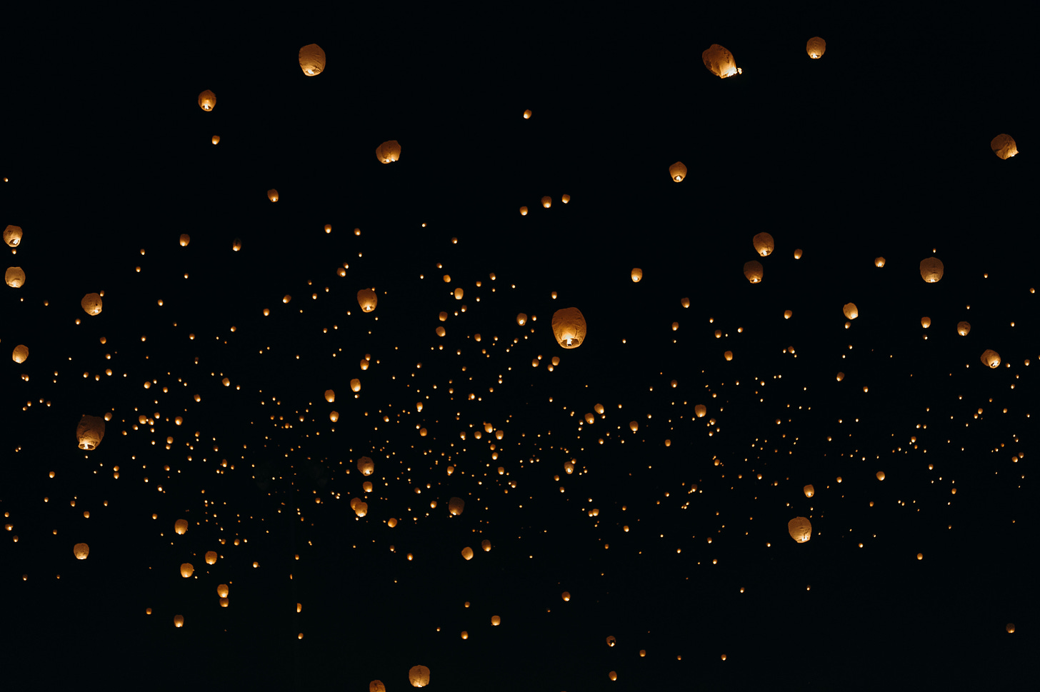 Photo of dozens of paper lanterns carrying lit candles, floating against a black background