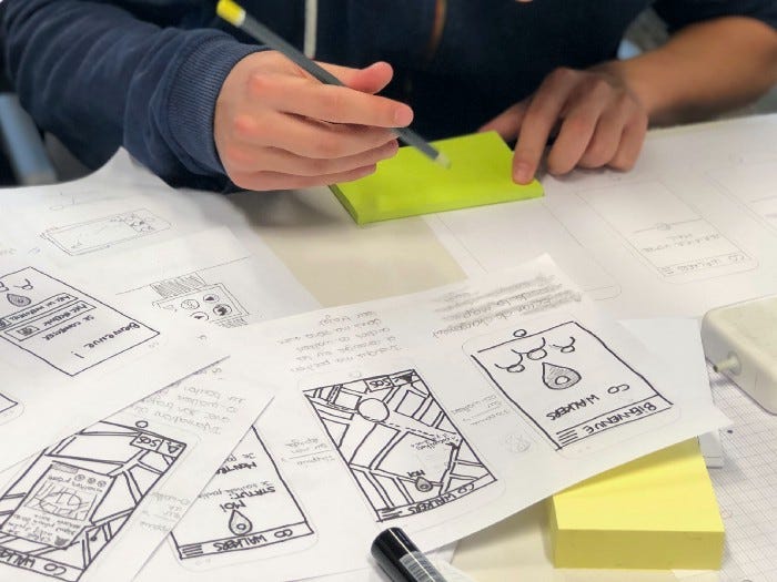 Paper sketches of a mobile app with a lot of text. The user is writing on a post-it note, and there is text below each paper screen and on the app.