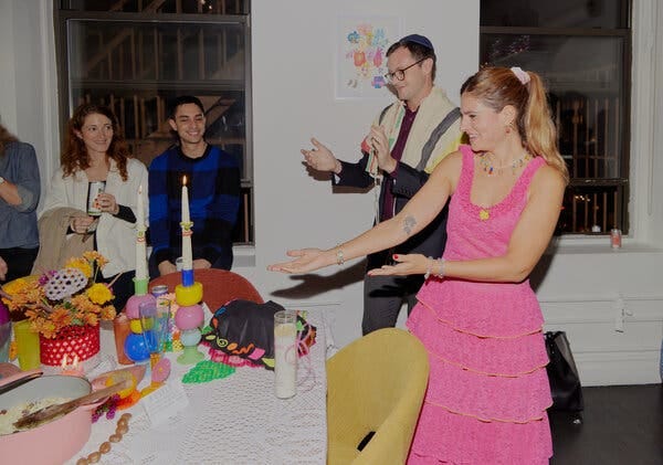 Susan Korn, wearing a tiered pink dress, gestures toward a Shabbat table while guests smile and Rabbi Matt Green applauds.