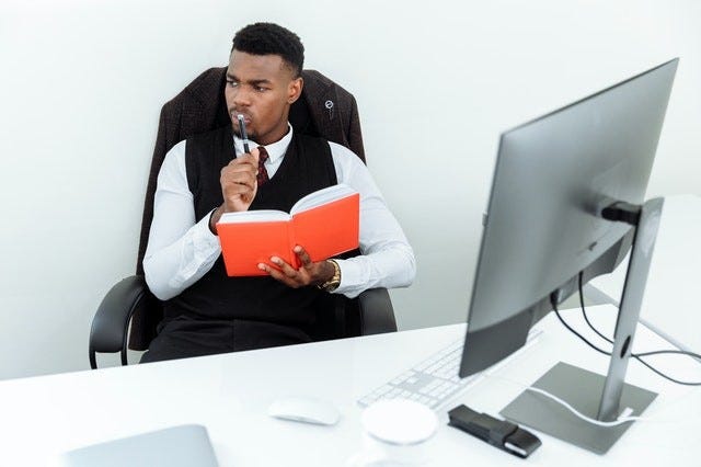 person sitting at a desk in fron of a desktop computer, holding a red notebook and thinking with a pen pressed to their lips