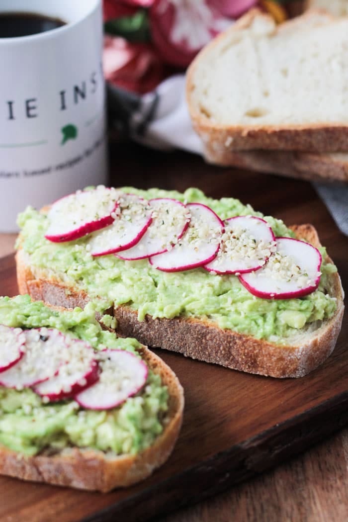 Two slices of avocado toast topped with sliced radishes and hemp seeds. Stack of bread in the background next to a mug of coffee.