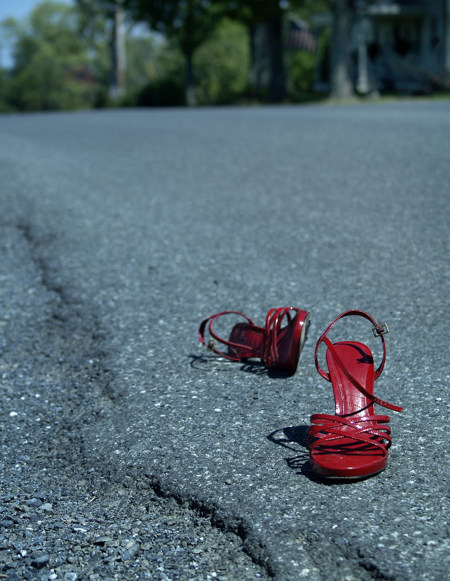 Red high heeled shoes with straps on the edge of an asphalt road. Very subtly in the background there is a small house flying an American flag