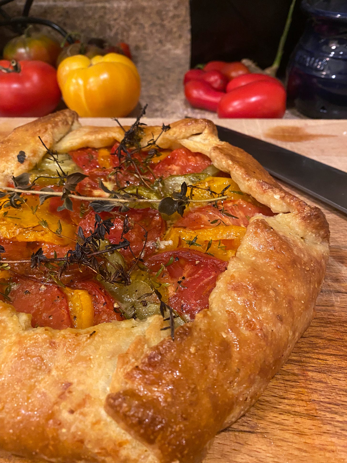 On a wooden cutting board, a golden brown crostata full of yellow, red, and green heirloom tomatoes. Sprigs of thyme, crisp from the oven, are scattered over the top. In the background a large knife rests on the board, and several whole heirloom tomatoes are visible.
