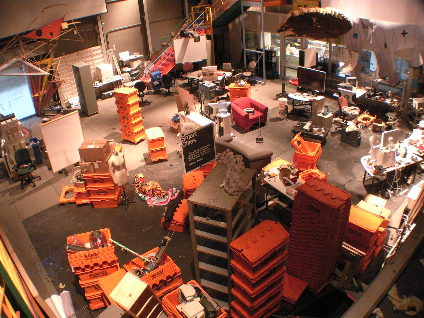 An aerial shot of the Smart Cities lab on a moving day to its current site. Orange crates, lab benches, storage unite, and odd scraps of furniture lay in disarray in an industrial space.