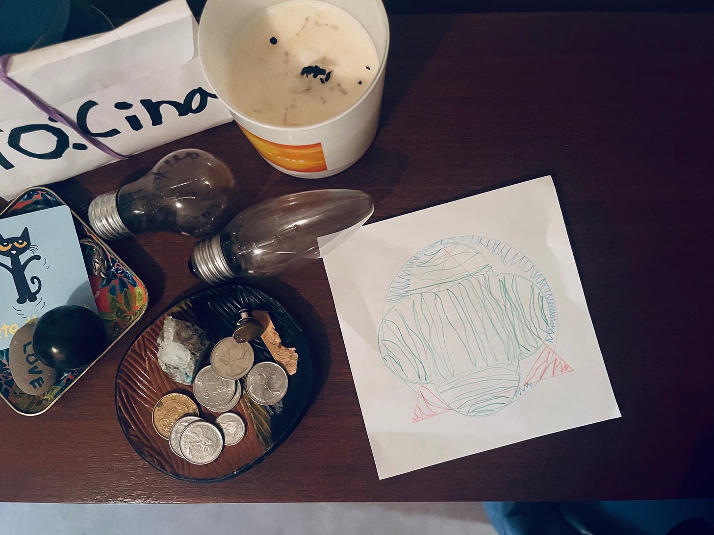 Aerial view of the sideboard that holds two blackened lightbulbs, a dirty candle, a pile of change, a Pete the cat tarot card, a rock on which is written “love” in a child’s handwriting, and a thick envelope that reads “to: cina” also in a child’s handwriting