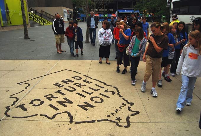 In an open plaza of tan brick, a large black stencil paints a contour of the state of Illinois, with the words "End Torture in Illinois" and a star near the southern tip of the state, and next to it the word "Tamms." A couple dozen people look at the image.