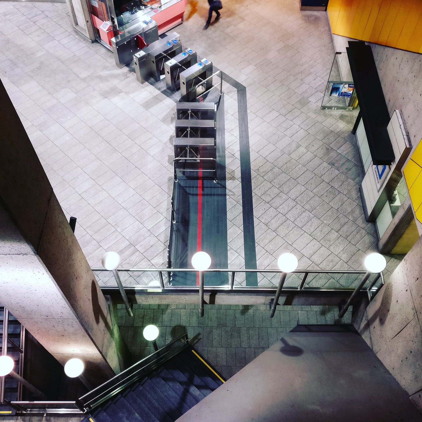 An empty subway station in Montreal, shot from the entrance, looking down at the turnstiles. The legs of a single person are visible at the top of the frame