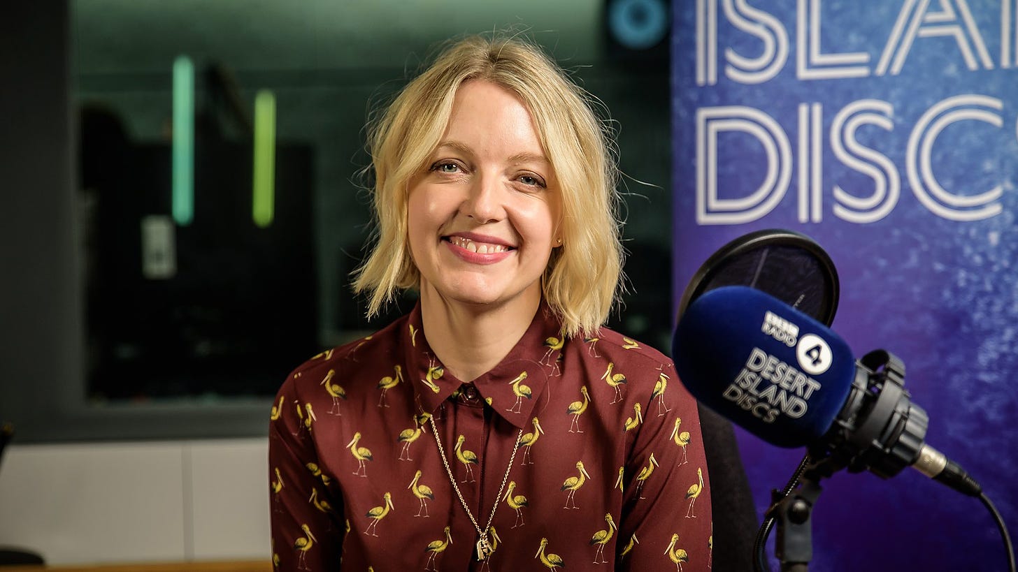 BBC Radio 4's Desert Island Discs host Lauren Laverne sits in front of a branded microphone the in the studio. Her blonde hair is centrally parted. She is wearing a brown shirt with pictures of storks on it. She is wearing a gold chain. 