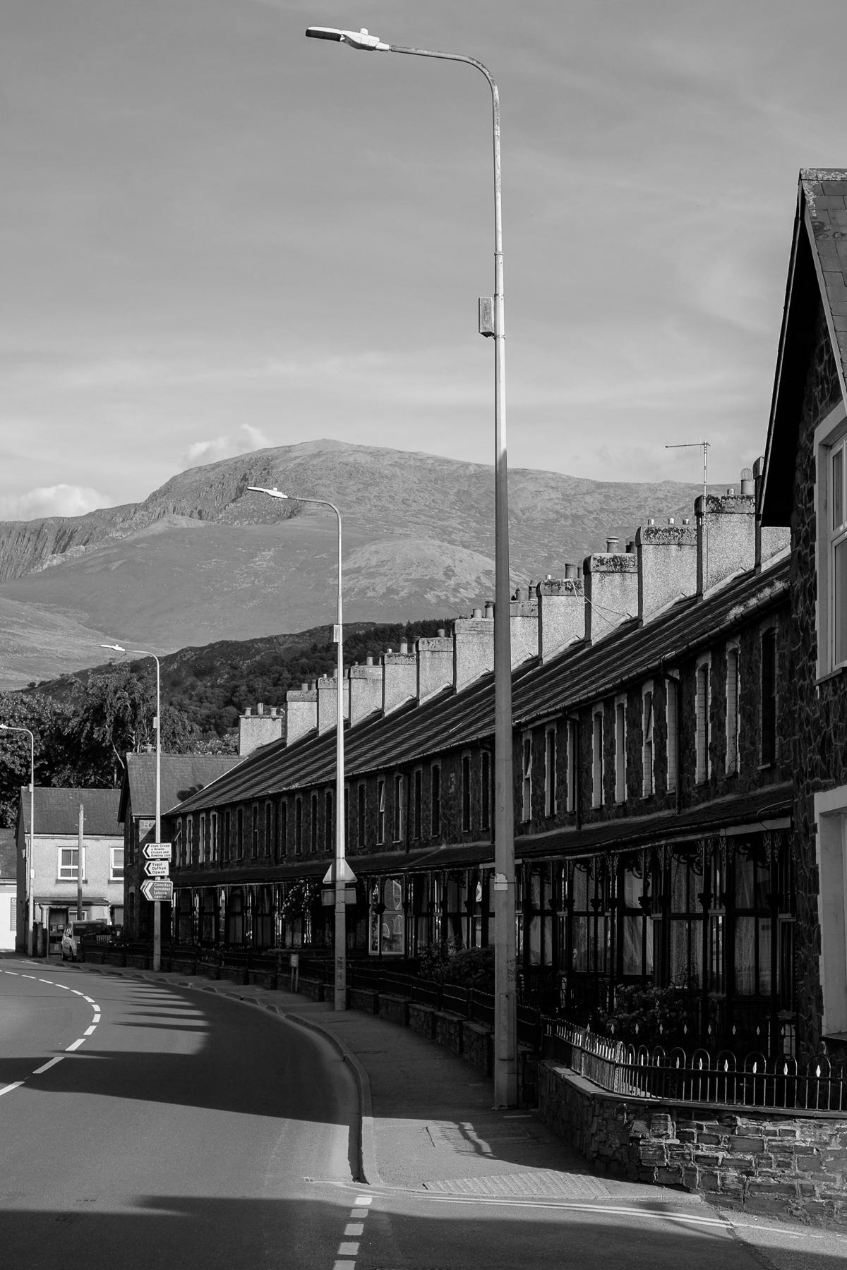 Black and white photograph of a row of terraced houses alongside a road with streetlights recedes into the distance towards a mountain.