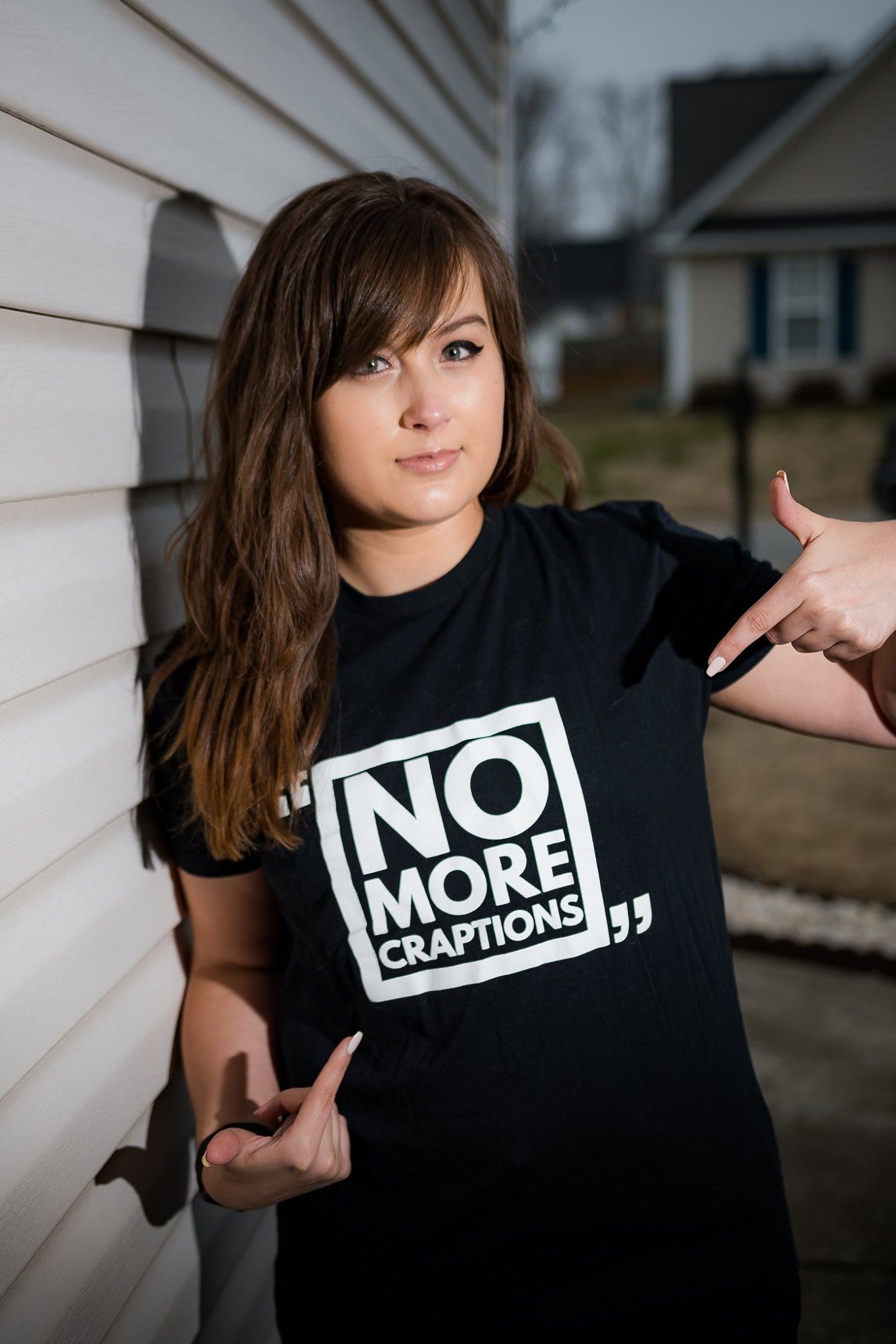 Rikki, a white woman with brown hair and bangs points at her black t-shirt, which says "No More CRAPtions" on it.