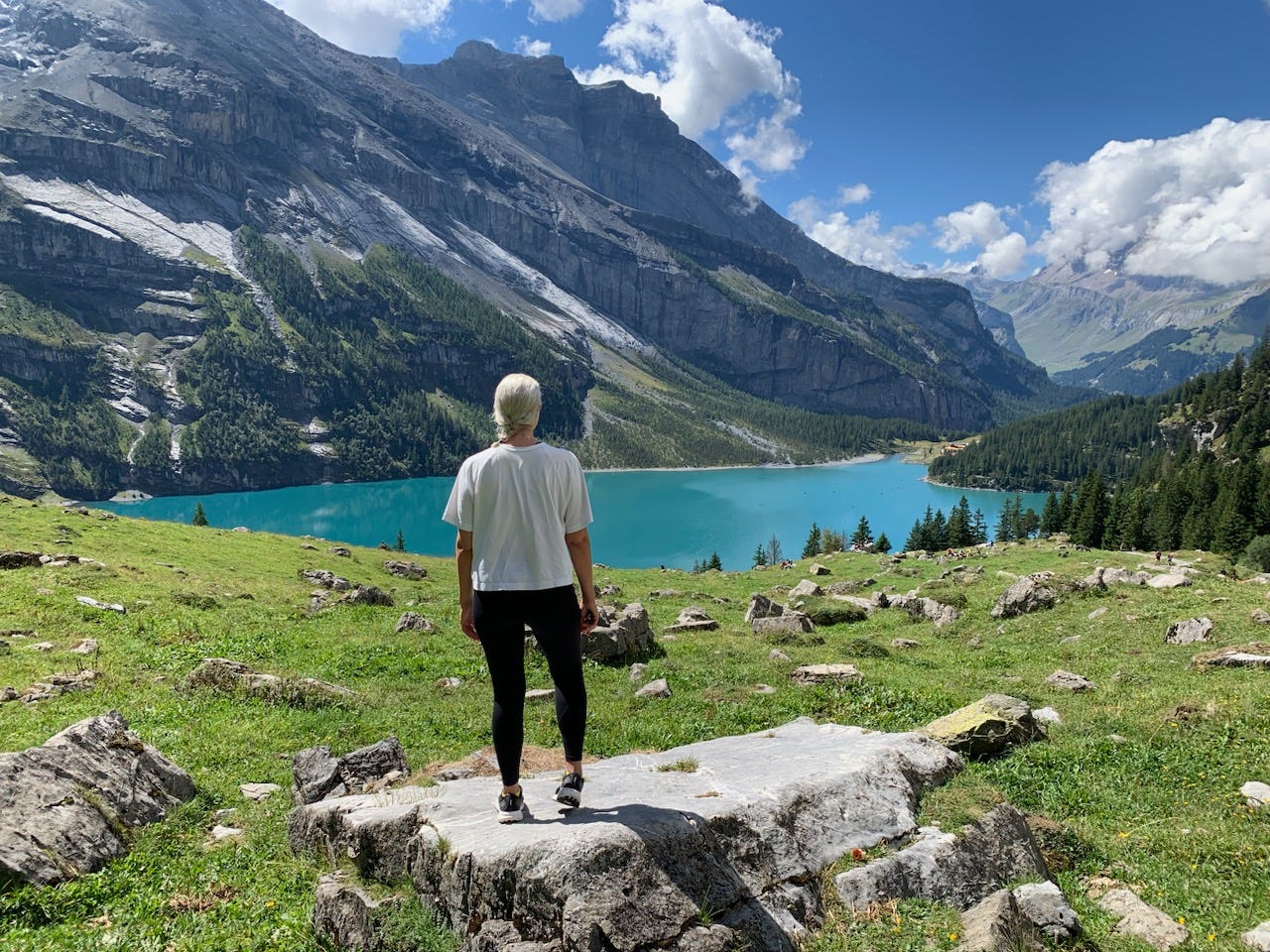 A blonde woman in a white T-shirt and black leggings stands with her back to the camera looking out on a picturesque view of mountains rising over an aqua blue lake.