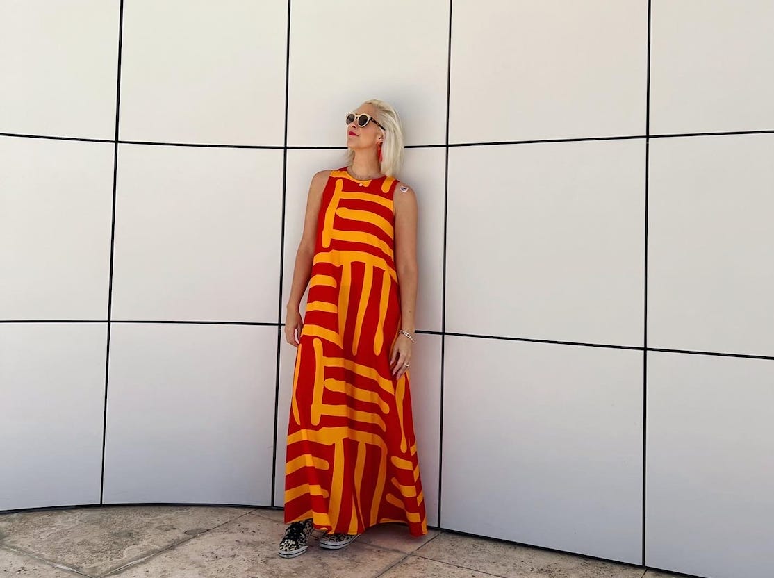 A blond women wearing gold sunglasses and a bold-patterned red and gold dress leans against a large, white-tiled wall.