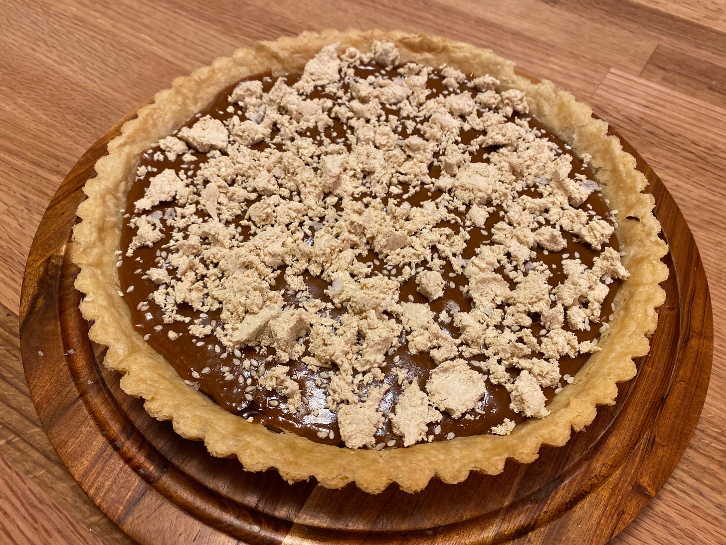 A round tart with fluted edges sits on a wooden platter. The chocolate filling is covered with crumbled pieces of halvah and sprinkled with sesame seeds.