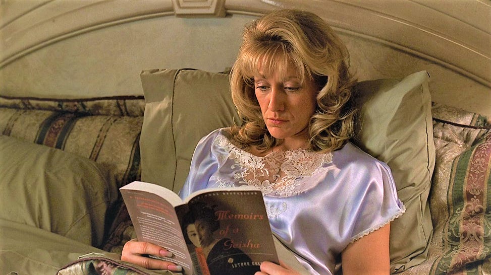 Carmela (personagem da série The Sopranos) deitada na cama, lendo o livro Memórias de uma gueixa.