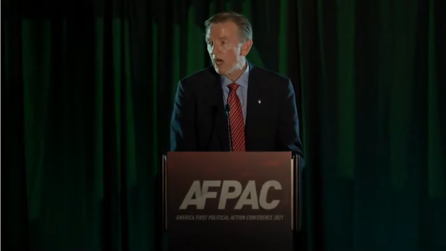 Paul Gosar is seen standing behind a lectern with the letters AFPAC on the front. He is standing in front of a dark green curtain. He is wearing a suit and tie, and his face is shiny with sweat.
