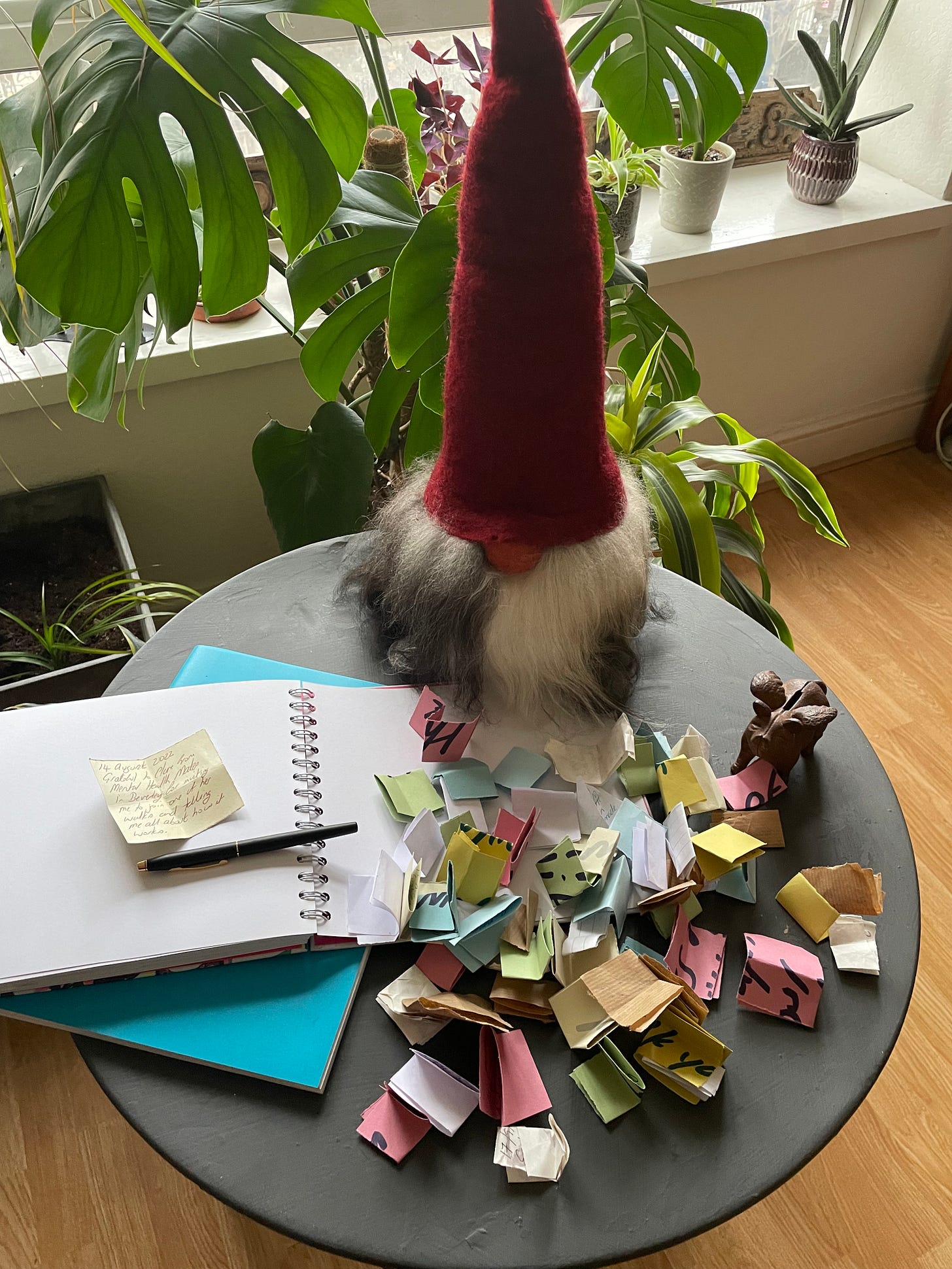 A table covered in folded scraps of coloured paper next to an open scrapbook with a pen. A stuffed Nordic gnome with a red hat is in the background.