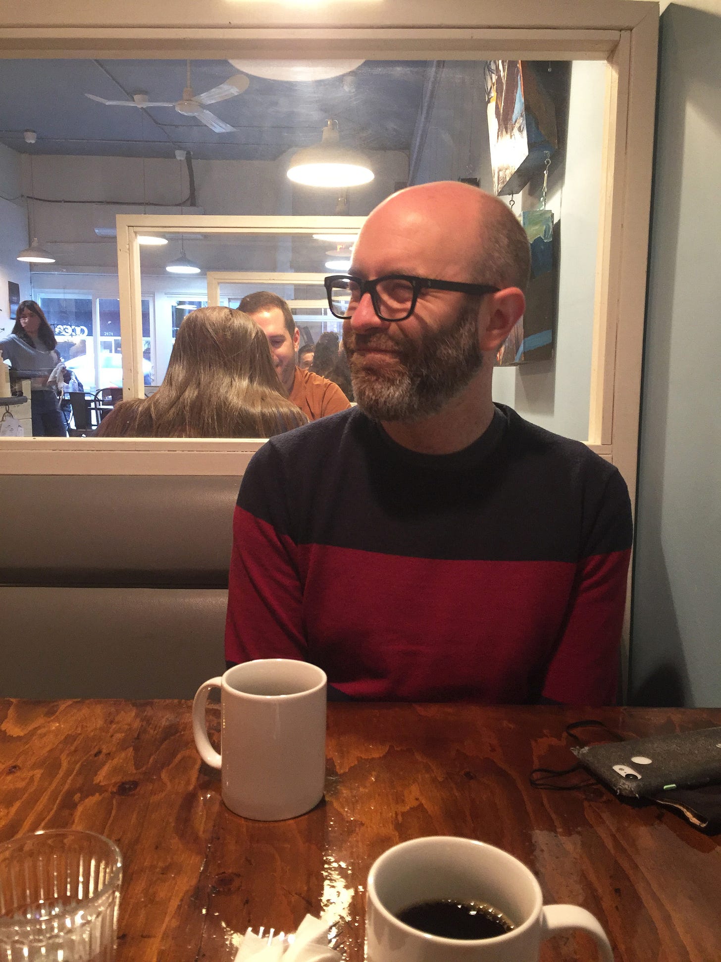 In a restaurant booth with two coffees on the table, Iain looks slightly away from the camera, smirking. He is wearing a blue and red colour blocked sweater.