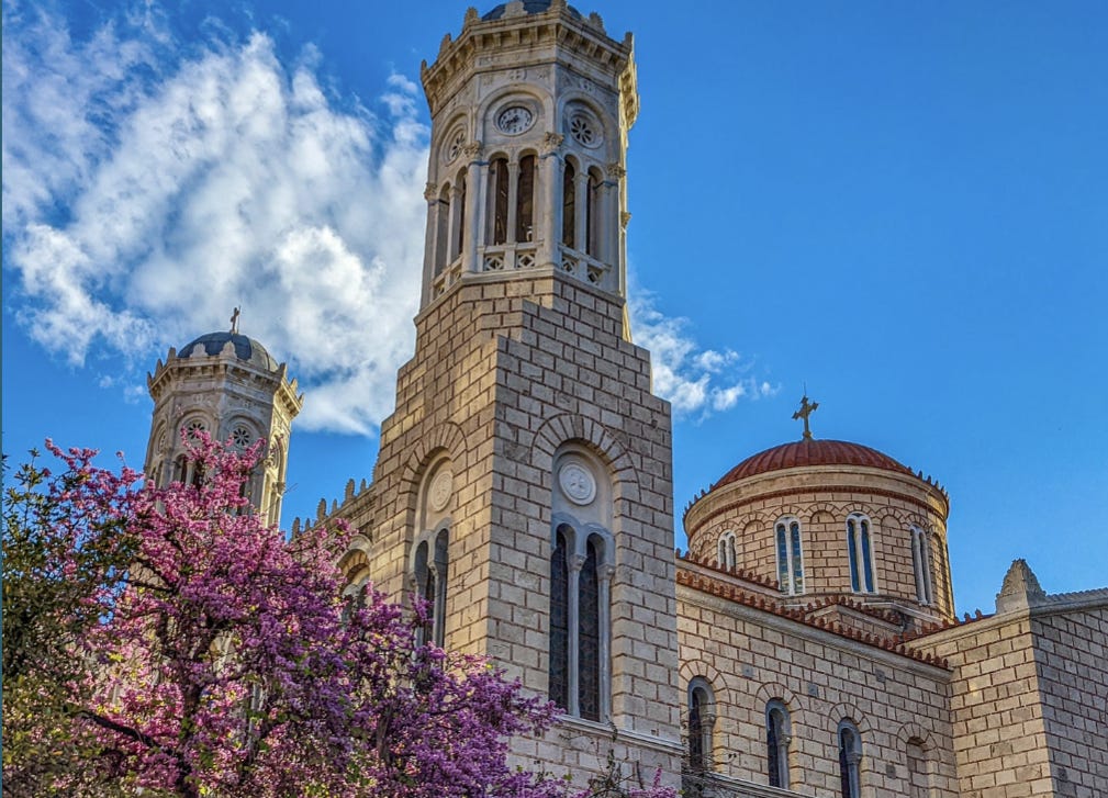 A photo showing the domes and tower of an Eastern Orthodox Church with a blue sky behind and a tree filled with purple blossoms in front. 