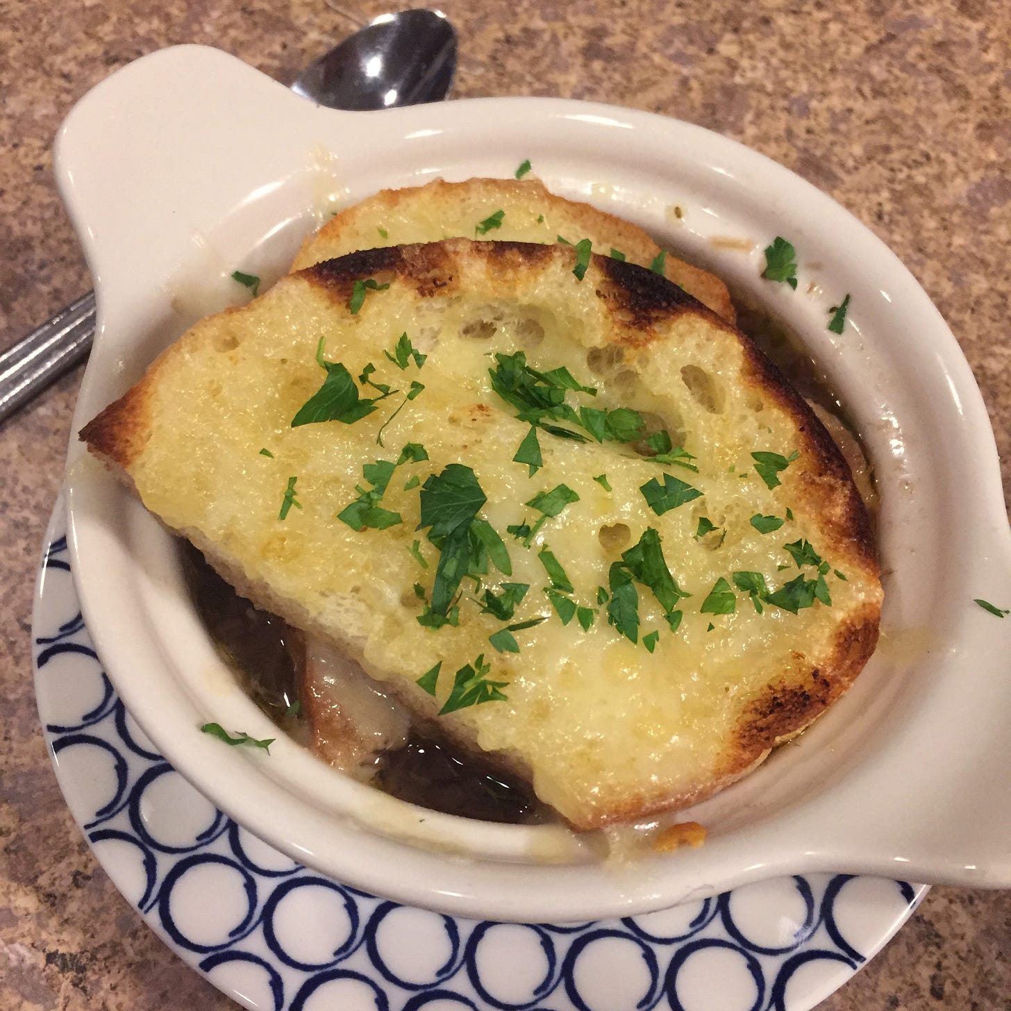 A white French onion soup bowl sits on a small blue and white plate with a spoon beside it. On top of the soup are two halves of a slice of bread, browned and with melted cheese, and a dusting of parsley.