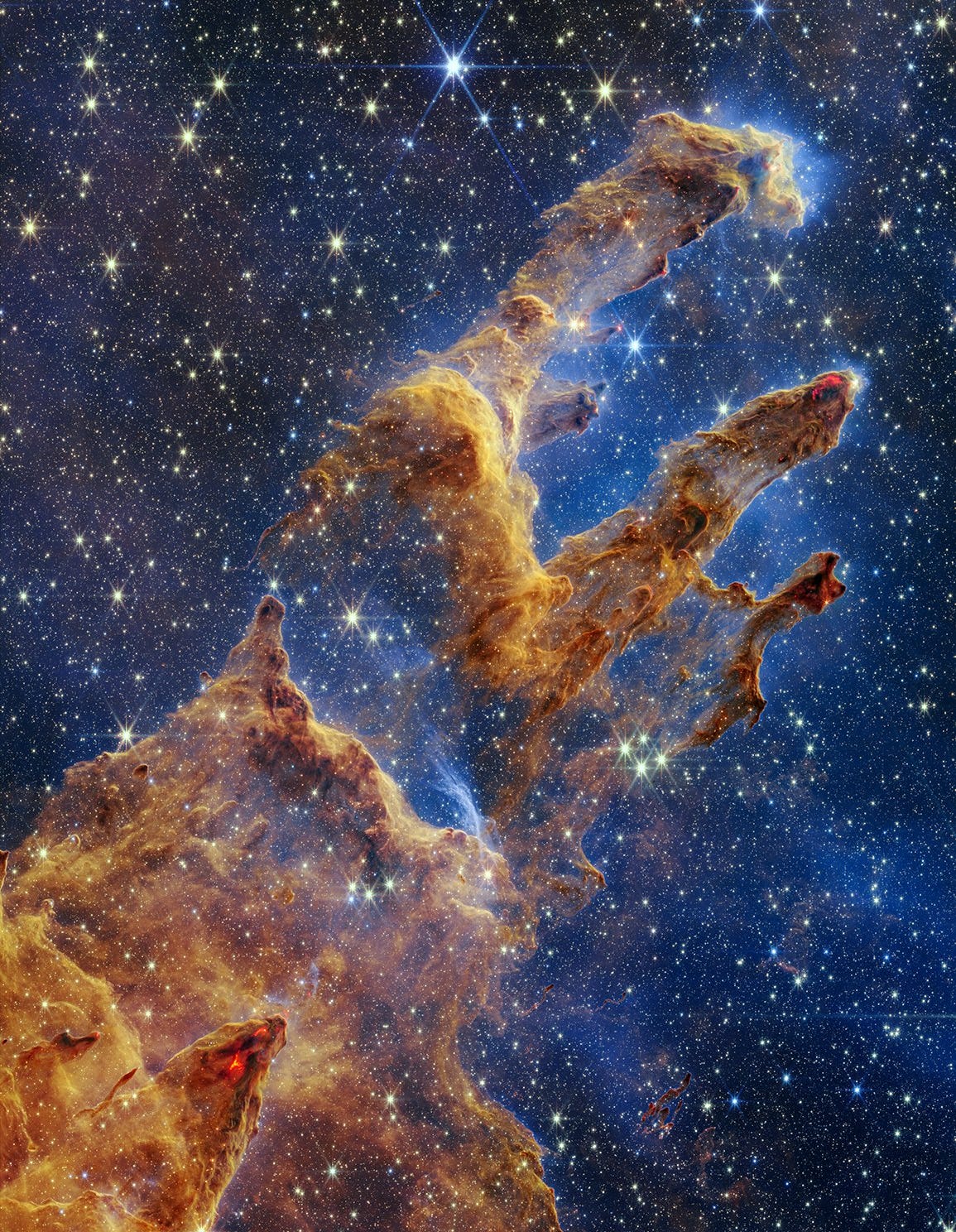 [inglise keeles] At the bottom left of this vertical image are the thickest regions of brown and rusty red gas and dust. There are many layers of semi-transparent gas and dust overlaying one another. A peak rises about a third of the way from the bottom, and becomes far darker brown with two bright red areas toward the tip. The light brown dust becomes more diaphanous about halfway up the screen. There’s a slight gap in the dust, which allows the blue background to come into view clearly. About 60% of the background in this image is set in shades of blue and littered with tiny yellow and blue stars. The brown pillars continue, taking the shape of a shoulder at the base, with three prominent columns rising out toward the upper right. The top left pillar is the largest and widest. The peaks of the second and third pillars are set off in darker shades of brown and have red outlines.