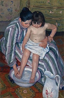 A mother dressed in a striped dress with long sleeves and turtleneck helps her small daughter wash her feet. She wears her shiny black hair in a loose bun and holds the child securely. The girl is sitting in her mother’s lap and her waist is wrapped in a white towel. She watches as her mother gently dips her small legs into the purple-rimmed glass bowl that stirs below with water. In the right-hand corner, a white water pitcher with different shades of pink flowers is standing on  the orange-patterned carpet. In the background a yellow wardrobe with tiny flowers and golden handles sits against the flowery wallpaper. 