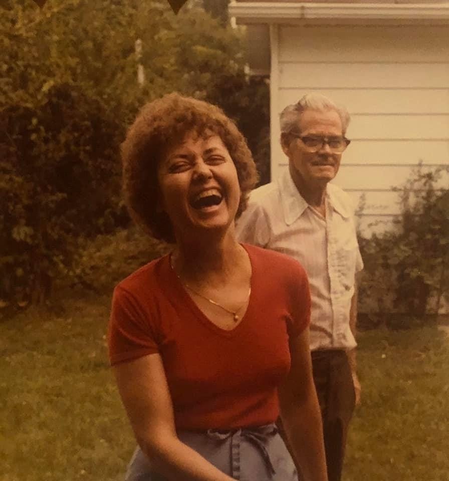 Setting is outside, green space in front of a white shed. Subjects are my grandmother, Nana, and great-grandfather, her dad. Nana is laughing with her mouth open and is wearing a small gold necklace, a red v-neck t-shirt, and baggy pants. The man behind her looks on, and is wearing a pin-striped button-down white-collar short-sleeve shirt and glasses. The photo is old-looking and taken in the 1970s.
