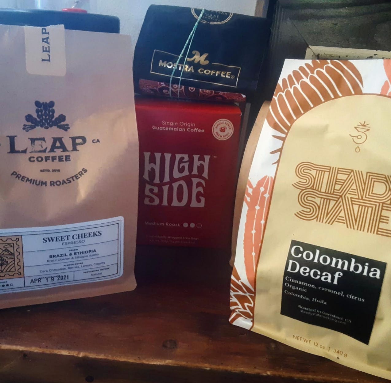 A bunch of different branded coffee bags on a worn wooden shelf.