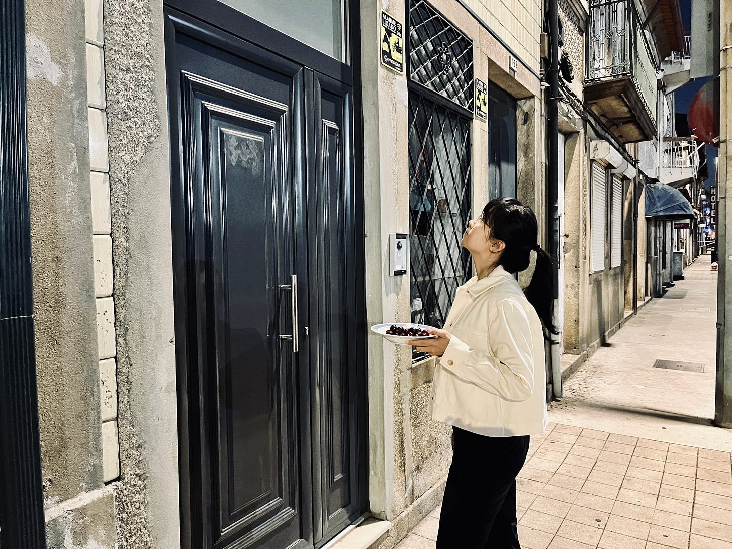 Image: Photo of me, at night, wearing a cream colour light summer jacket, hair tied up to a ponytail, holding a plate of cherries, looking upwards to my neighbour’s door, waiting for them to open the door for my husband and me. The background shows a curving row of shops and houses alongside a road with streetlights. 