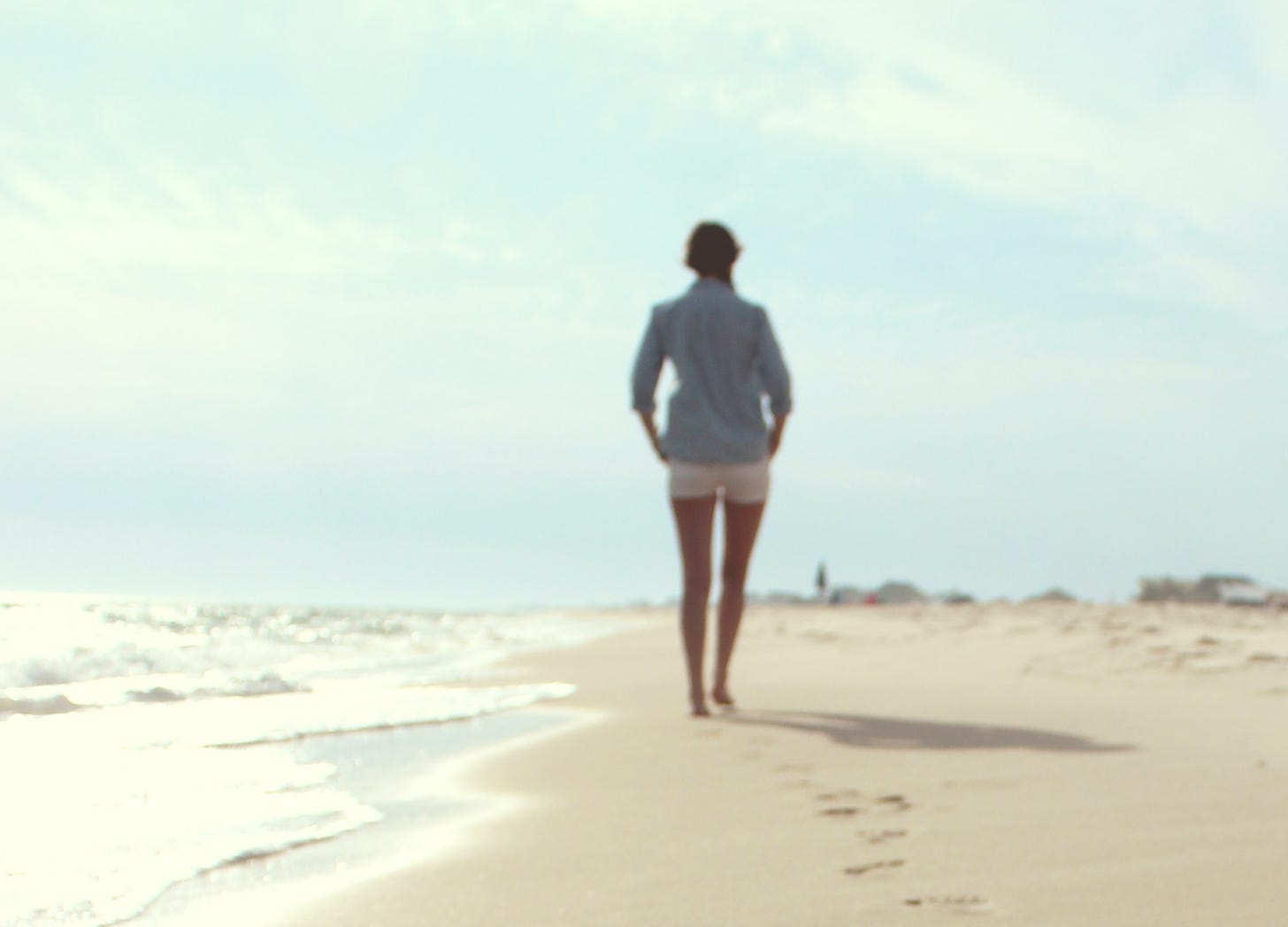 Mulher de short branco e camisa azul clara de mangas compridas dobradas caminha descalça em areia da praia com o mar quebrando as ondas do seu lado esquerdo, em dia de céu azul.