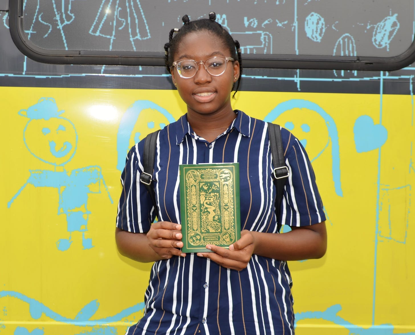 A young black writer with short braided hair, clear glasses, and a dark blue shirt with white stripes. She stands in front of the ABC mobile holding the ABC anthology, Pandemic’s Labyrinth.
