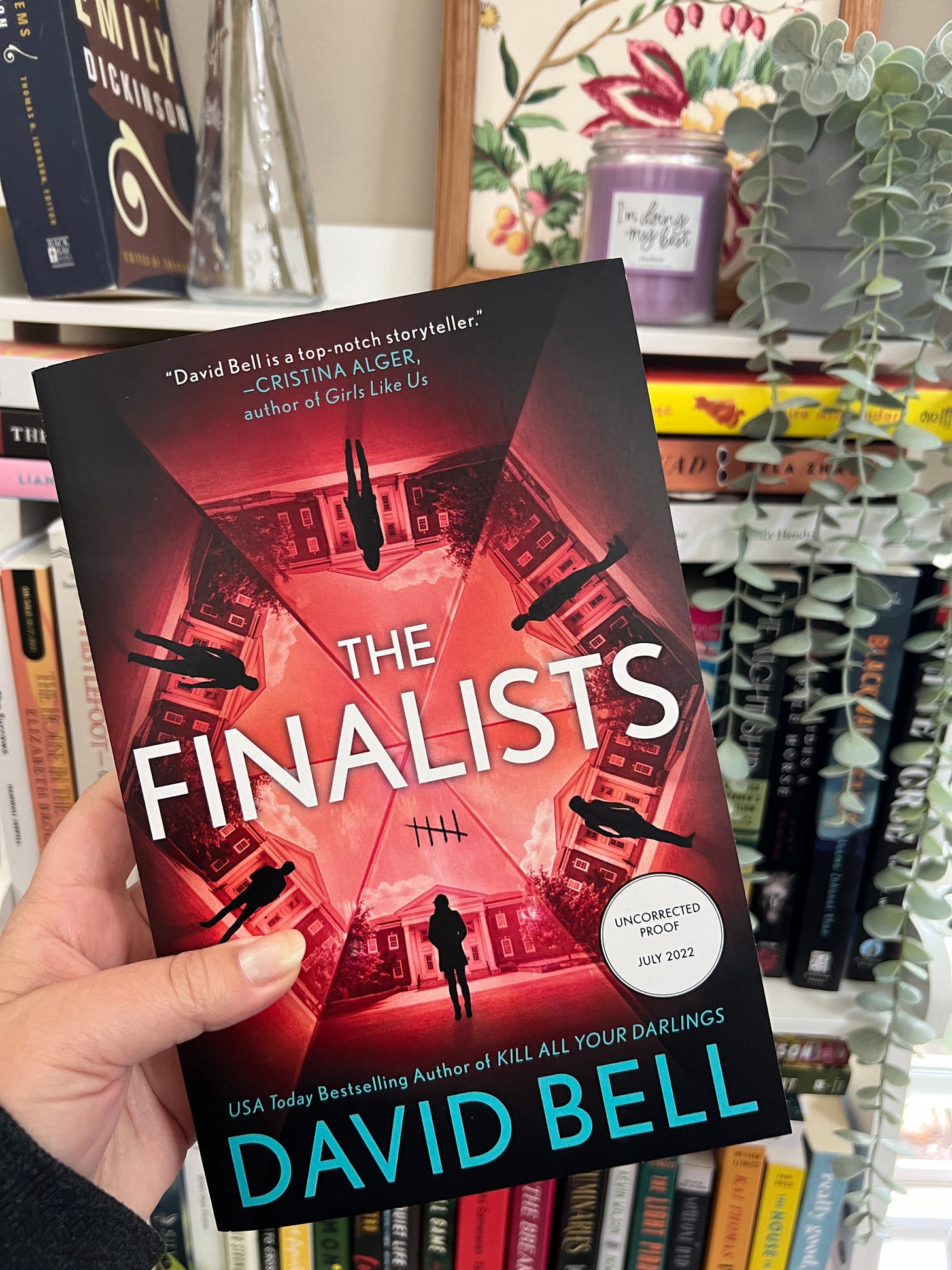 A white hand holding an ARC of The Finalists up in front of a full bookshelf. 