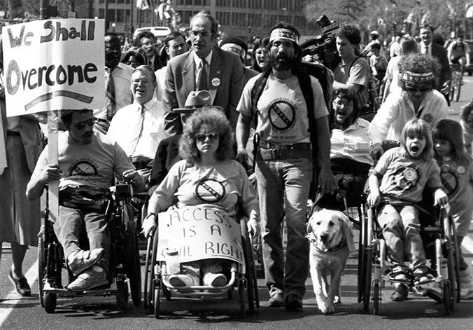 A black and white photo of a group of people marching. Some people are using wheelchairs and some are walking. They are holding signs that say "We shall overcome" and "Access is a civil right."