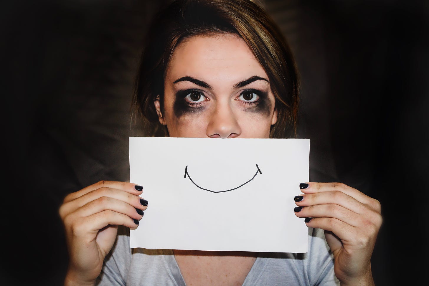 Photo of a woman with black eye makeup holding up a paper with a smiling face over her mouth. Sydney Sims / Unsplash