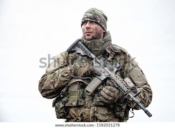 Portrait of soldier, modern combatant with dirty face, firearm replica, wearing camouflage uniform, beanie and masking cape on neck, standing on field, carrying backpack on march