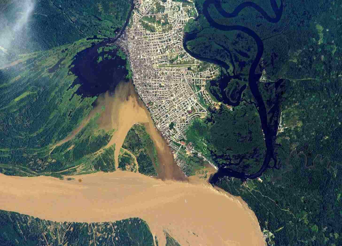 A satllite image of the grey streets of the city of Iquitos on the edge of waterways flowing into a huge brown river, surrounded by green forest.