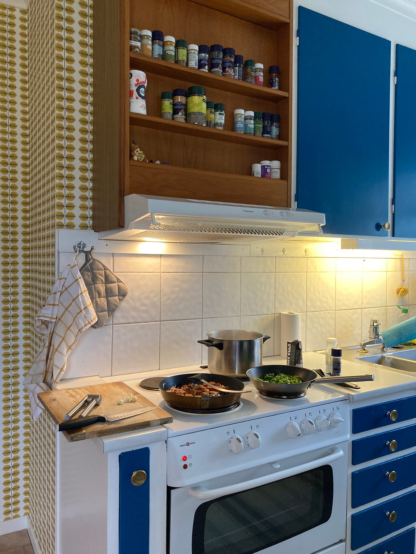 A kitchen with blue cupboard doors, yellow wallpaper, and a wooden spice shelf above a white stove with pans on it.