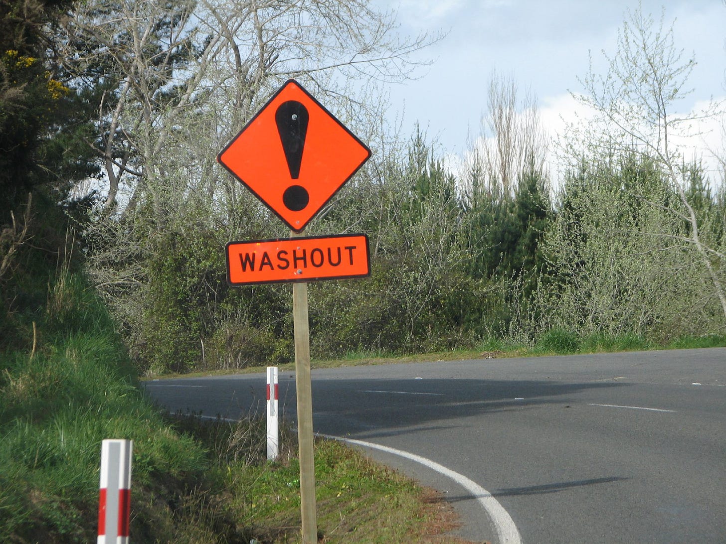 Curving hillside road with a bright red sign showing an exclamation mark and the word WASHOUT.